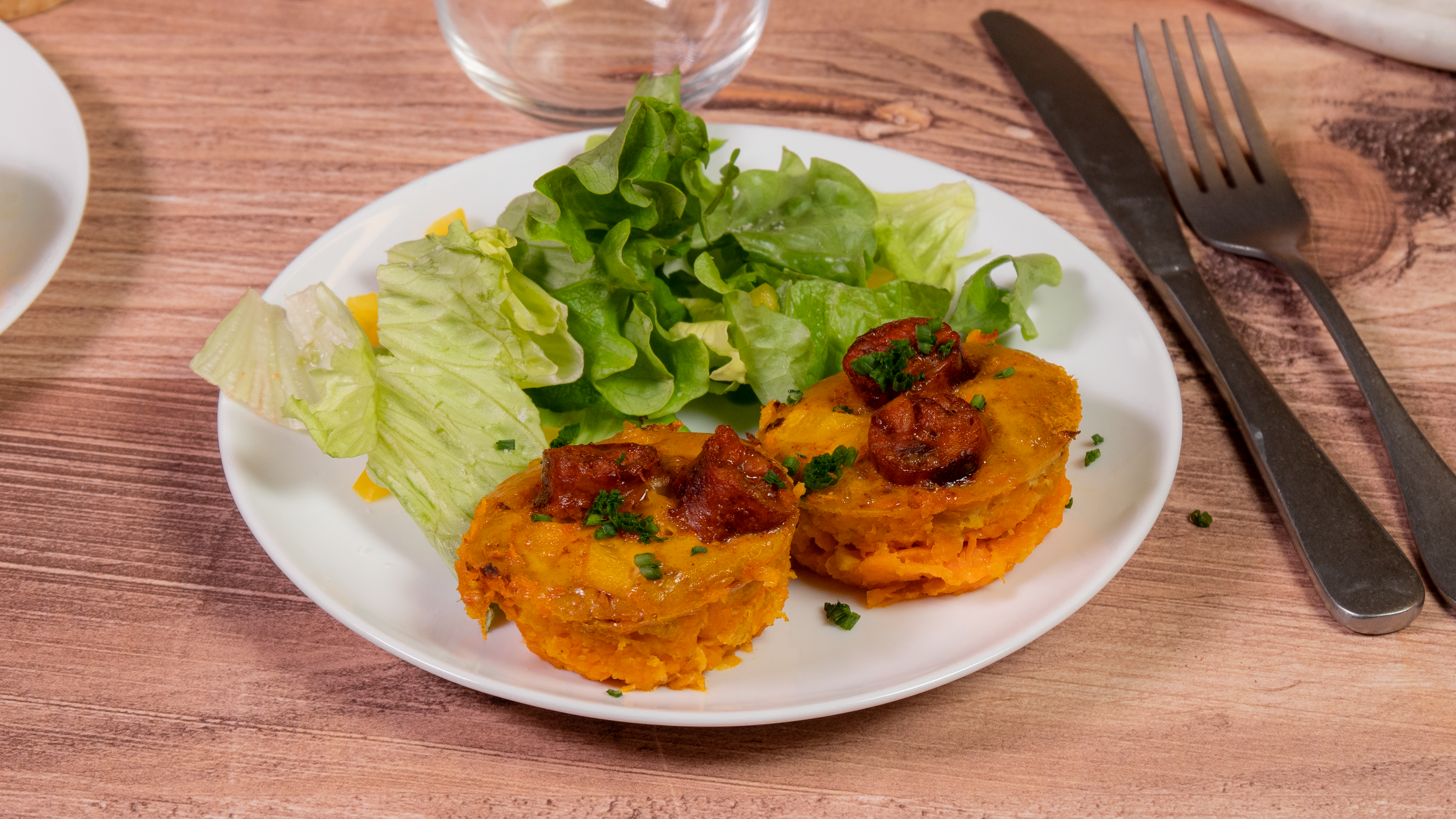 Rôti aux saucisses de volaille saveur chorizo Réghalal dans une assiette et accompagné de salade. L'assiette est posée sur une table en bois, avec des couverts et un verre